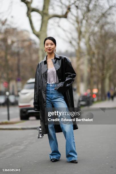 Guest wears gold large chain pendant earrings, a black shiny leather oversized long coat, high waist blue faded flared pants, a silver nailed /...