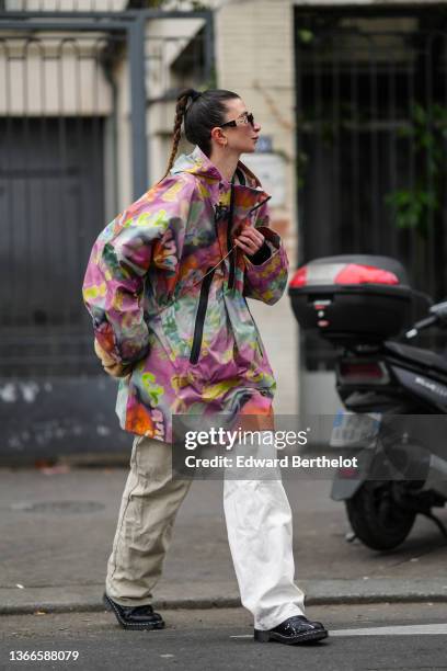 Guest wears black sunglasses from Versace, a purple / pale green / orange / neon yellow print pattern hoodie / high neck oversized rain coat, white...