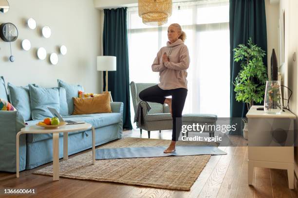 mature woman doing her morning yoga exercises in the living room - tree position stock pictures, royalty-free photos & images
