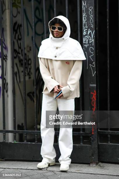 Guest wears beige sunglasses, a white denim hoodie buttoned shoulder cloak, a beige wit red logo inscription sweater from Wooyoungmi, white large...