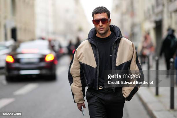 Guest wears red sunglasses, a black sweater, a black and beige print pattern oversized bomber coat, black suit pants, a black leather belt, outside...