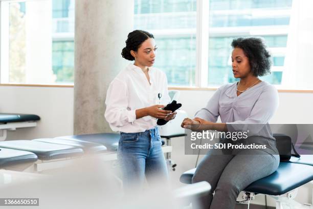 geschäftsfrau, die am untersuchungstisch sitzt, bespricht verletzung mit ergotherapeutin - physiotherapie praxis stock-fotos und bilder