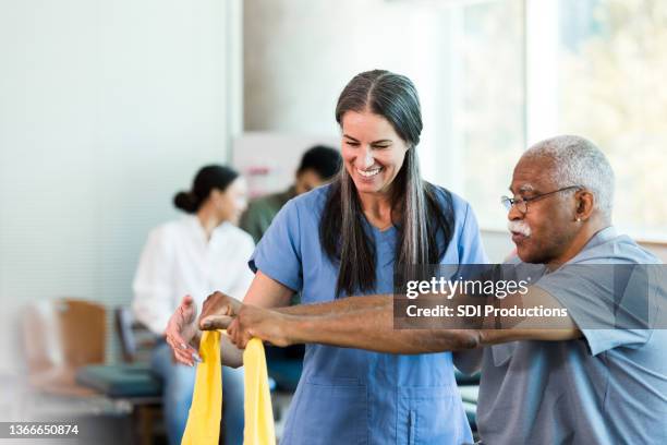 therapist smiles as senior man learns to use elastic band - fysiotherapie stockfoto's en -beelden