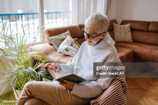 senior woman reads a book in the living room - literature stock pictures, royalty-free photos & images