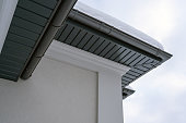 Corner of the house with new gray metal tile roof and rain gutter covered by snow at winter. Metallic Guttering System, Guttering and Drainage Pipe Exterior
