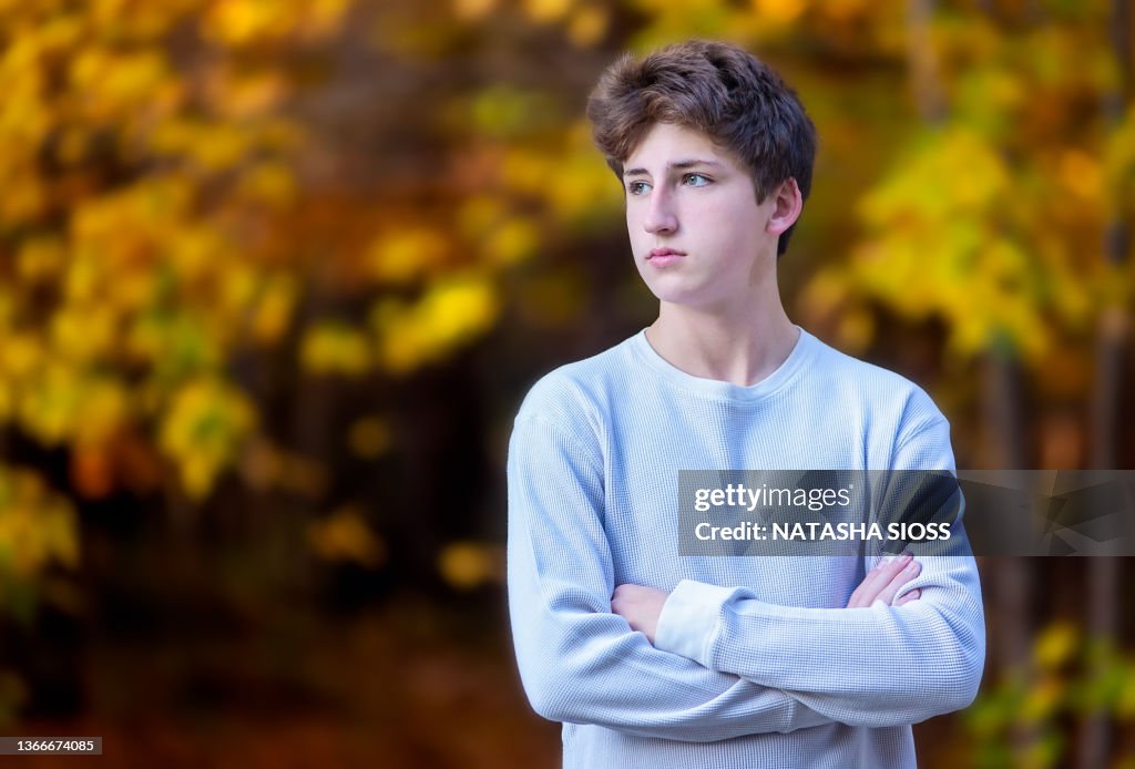 Teenage boy with brown hair and hazel eyes standing with arms crossed outdoors