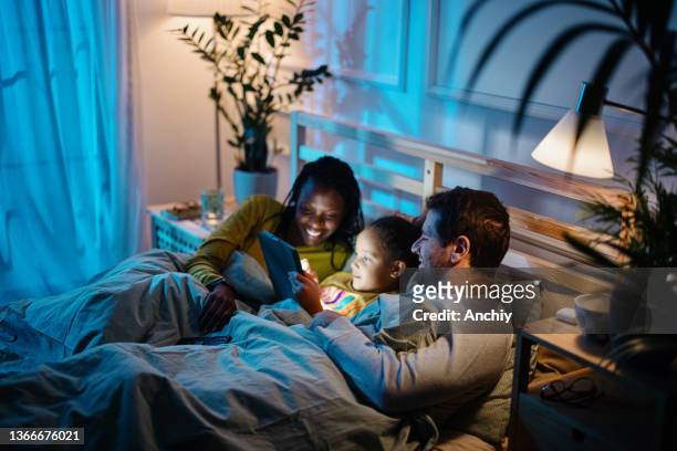 multiethnic family using a digital tablet together in bed at night. - mother happy reading tablet stock pictures, royalty-free photos & images