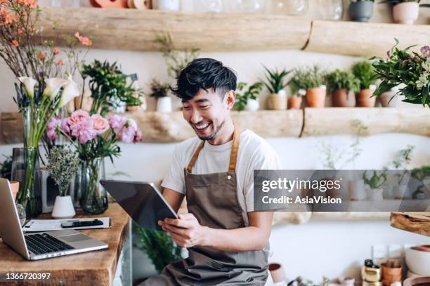fleuriste asiatique, propriétaire d’un magasin de fleurs de petite entreprise, utilisant une tablette numérique tout en travaillant sur un ordinateur portable contre les fleurs et les plantes. vérification des stocks, prise des commandes des clients, - auto entrepreneur photos et images de collection