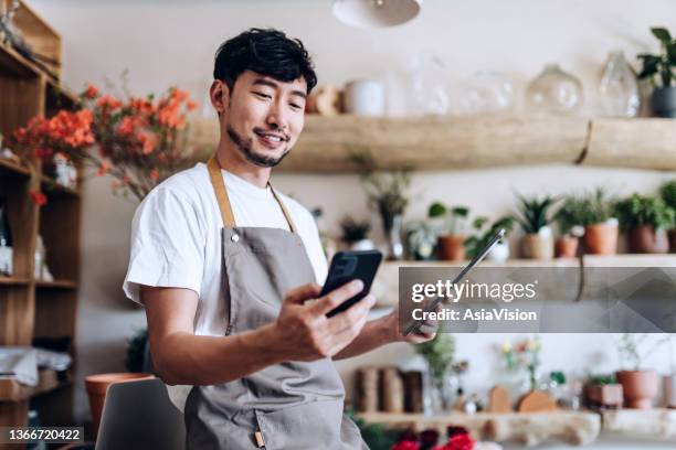 fleuriste asiatique, propriétaire d’un magasin de fleurs pour petites entreprises, utilisant un smartphone tout en travaillant sur une tablette numérique sur le lieu de travail. vérification des stocks, prise des commandes des clients, vente de produi - tablier photos et images de collection