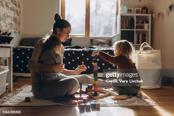 young mother playing with children while sitting on floor at home with wooden toys - mums stock-fotos und bilder