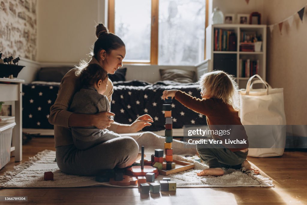 Young Mother playing with children while sitting on floor at home with wooden toys