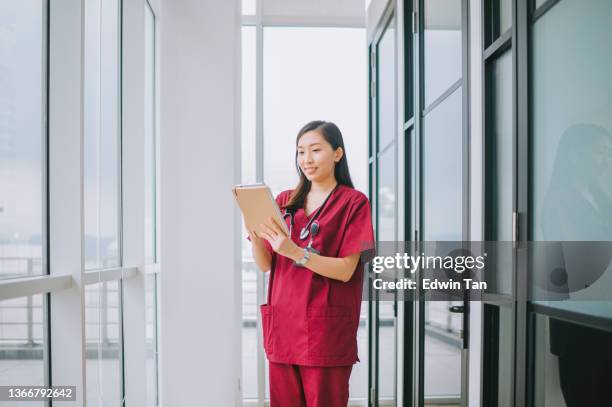 chinese female nurse healthcare taker smiling looking at clipboard on patient's health report in hospital corridor - ziekenhuiskleren stockfoto's en -beelden