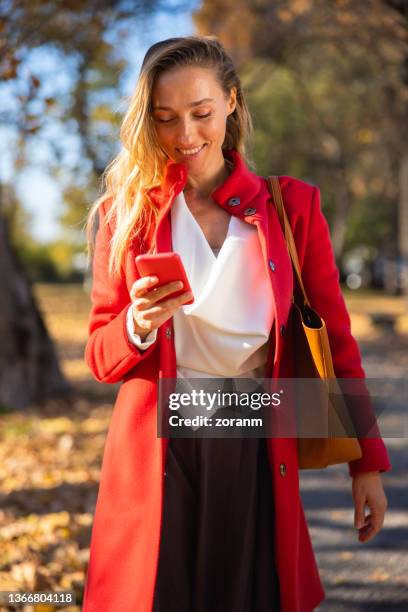 elegant woman smiling and waking in the park and using digital banking app on her smart phone - red billed stock pictures, royalty-free photos & images