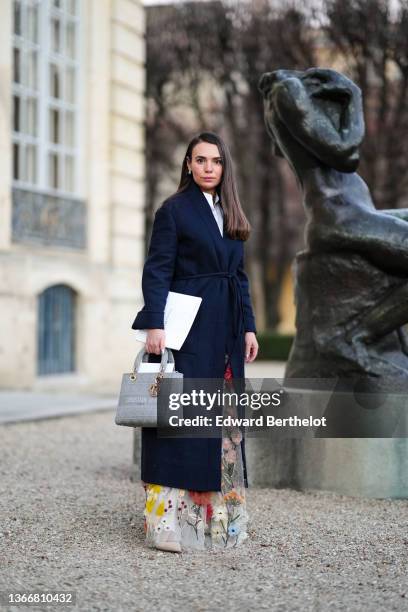 Guest wears a silver flower pendant earrings, a white shirt, a navy blue belted long coat, a white tulle with multicolored embroidered flower pattern...