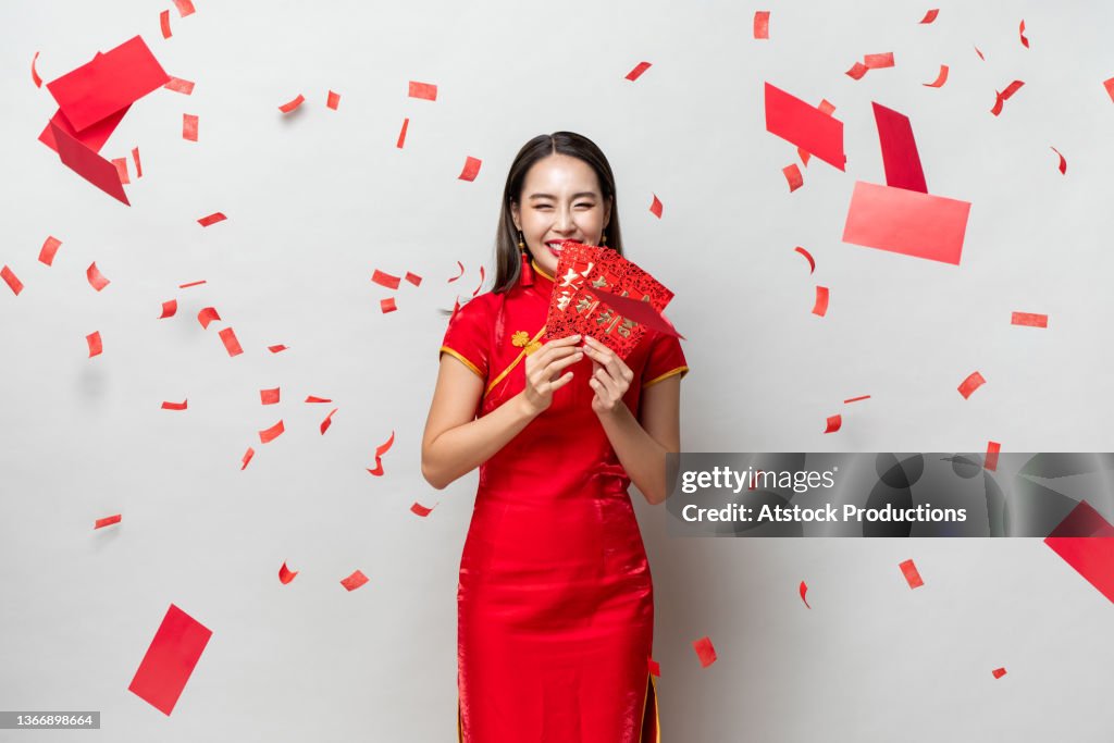 Smiling happy Asian woman in traditional oriental costume holding red envelopes Ang Pow in gray background, Chinese text means great luck great profit and and everything goes well