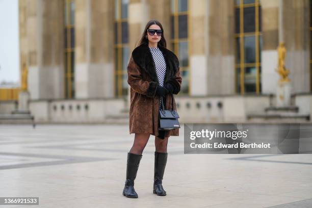 Bengi Guzel wears black futurist sunglasses, silver earrings, a black and white checkered print pattern high neck tube short dress, a brown suede...