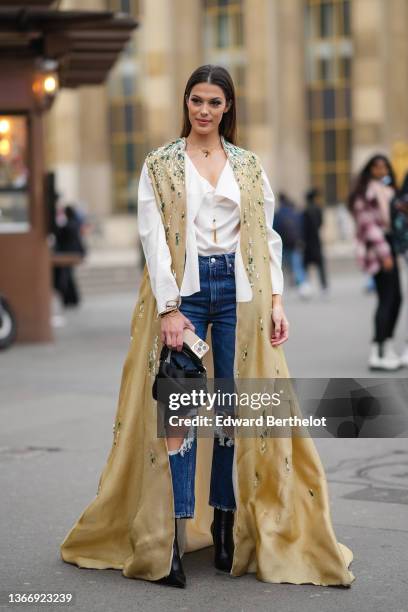 Iris Mittenaere wears a gold large chain necklace, a white oversized silk shirt, a brown with embroidered green and white pearl pattern long...