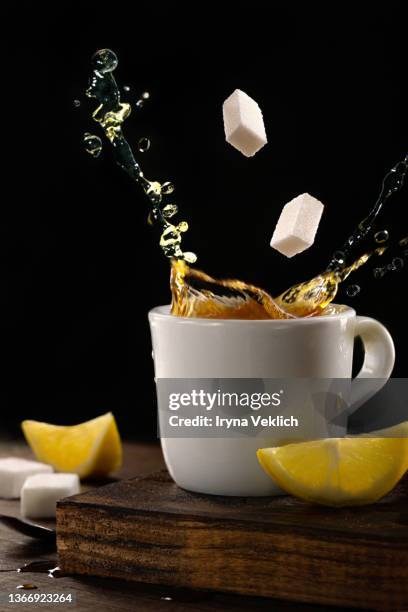 white mug with tea and splashes with white cubes of sugar, slices of lemon on dark black background. - sugar cubes in tea stock pictures, royalty-free photos & images