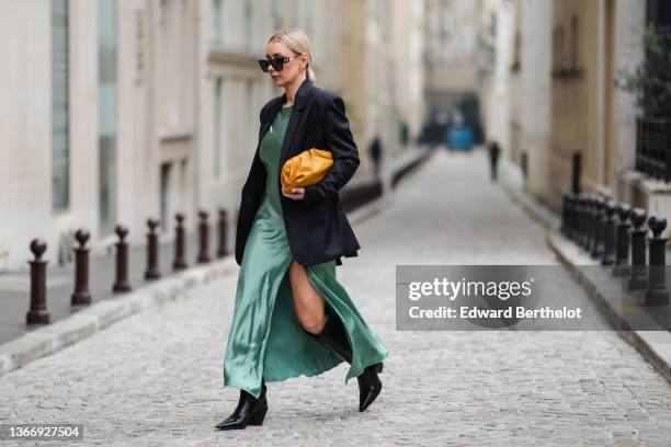Justyna Czernia wears sunglasses, gold earrings, a green mint satin silk slit / split long dress, a black oversized blazer jacket, an orange shiny...