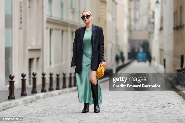 Justyna Czernia wears sunglasses, gold earrings, a green mint satin silk slit / split long dress, a black oversized blazer jacket, an orange shiny...