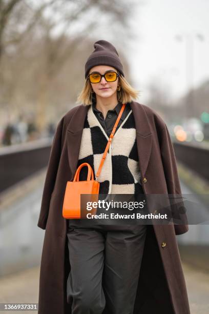 Guest wears gold earrings, a brown ribbed wool beanie, orange sunglasses, a black shirt, a black and beige checkered print pattern fluffy jacket, a...