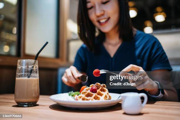 beautiful smiling young asian woman eating waffle with fresh cream and strawberries and having coffee in cafe - waffle stock-fotos und bilder