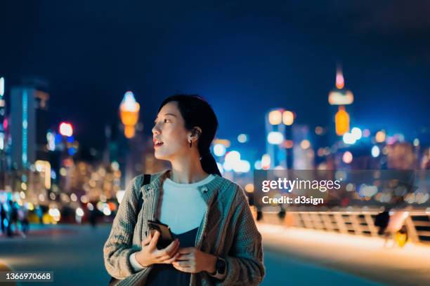 young asian woman using smartphone while walking along the promenade in the city at night, looking sideways, with illuminated and multi-coloured city skyline in background. lifestyle and technology - asia skyline stock pictures, royalty-free photos & images