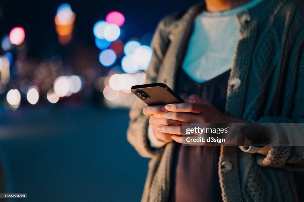 Close up, mid-section of young Asian woman using smartphone in illuminated and multi-coloured city at night. Lifestyle and technology