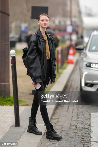 Model wears a brown and gold fluffy wool, a black shiny leather oversized jacket, a black shiny leather back pack bag, black skinny denim jeans...