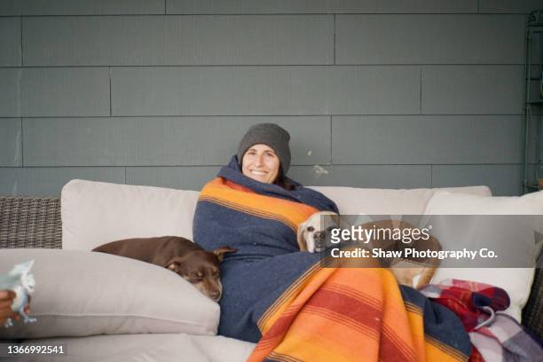 a woman bundled up in a blanket and hat on an outdoor couch with her two dogs snuggled into her. she is smling and looking at the camera - wrapped in a blanket stockfoto's en -beelden