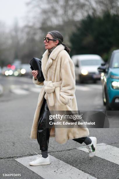 Guest wears black glasses, a beige / brown / yellow print pattern scarf, a white fur long belted coat, black shiny leather large pants, a black shiny...