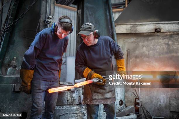 engineer with apprentice inspecting a forged steel part in industrial forge - smederij stockfoto's en -beelden
