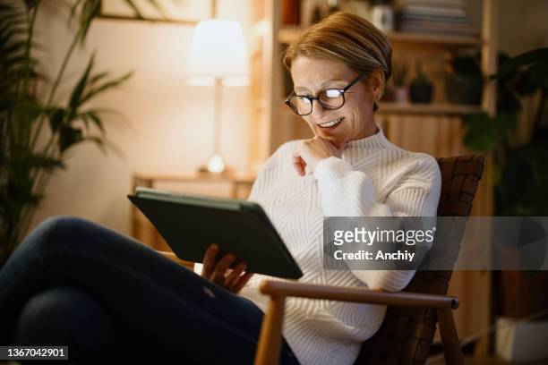 a mature woman reads on a digital tablet at night at home and laughs - vrouw 50 jaar stockfoto's en -beelden