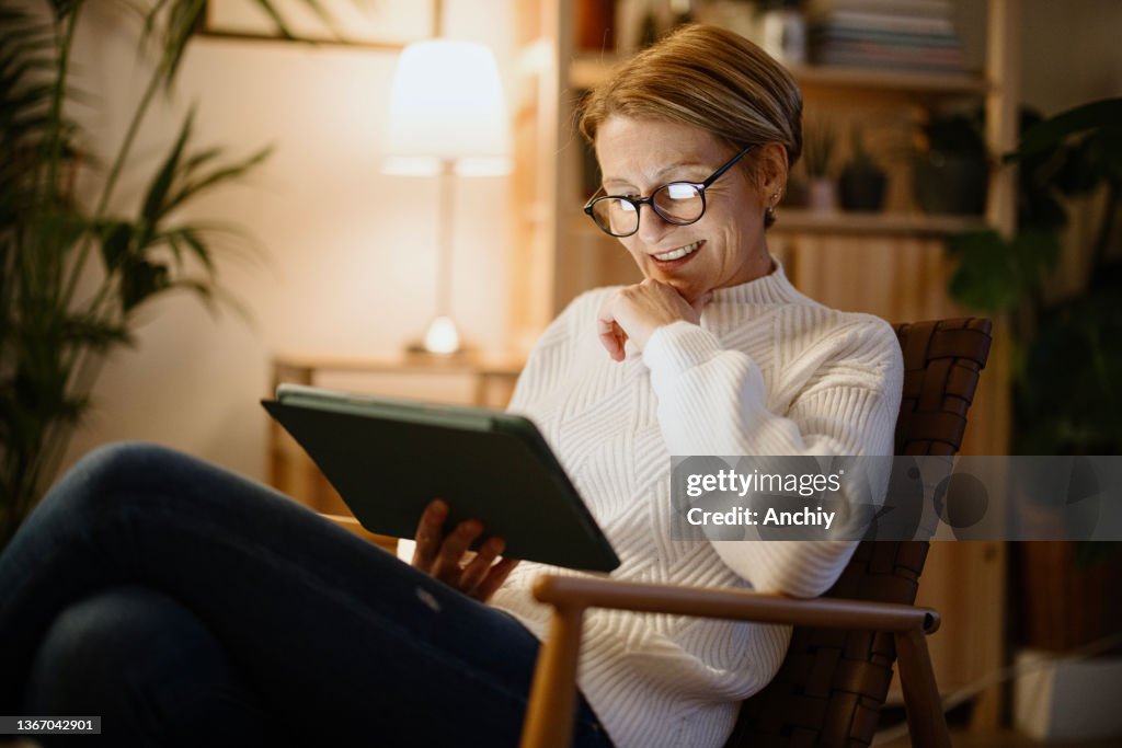 A mature woman reads on a digital tablet at night at home and laughs