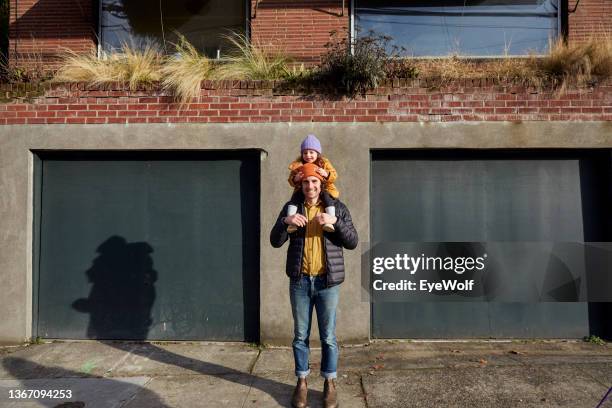 a young father walking around city with his daughter sitting on his shoulders, looking into camera smiling. - mann mit kind auf den schultern stock-fotos und bilder