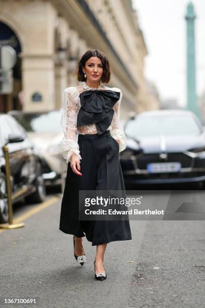 Alexandra Pereira wears large pearl pendant earrings, a white lace print pattern shoulder-pad shirt, a black oversized tie-bow, a black high waist...