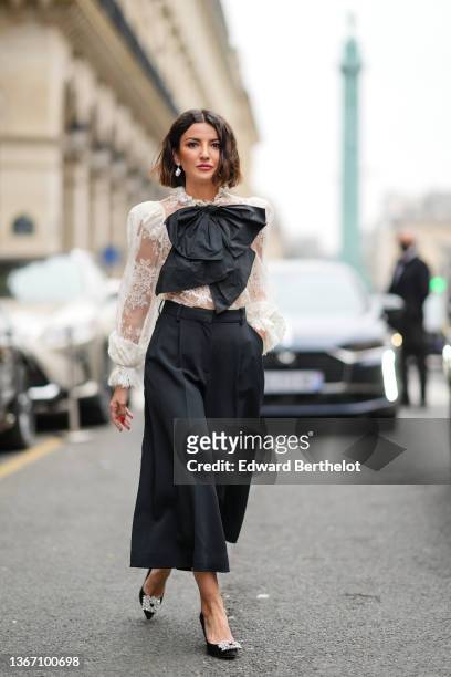 Alexandra Pereira wears large pearl pendant earrings, a white lace print pattern shoulder-pad shirt, a black oversized tie-bow, a black high waist...