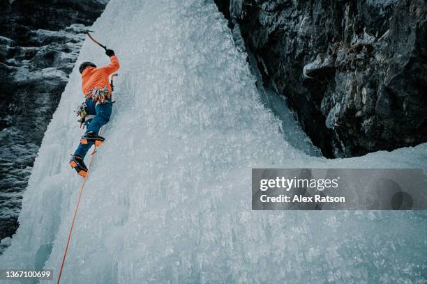 low angle perspective of a male ice climber swinging his ice axe into a tall frozen waterfall - extreme sports point of view stock pictures, royalty-free photos & images