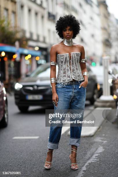 Didi Stone wears a silver large necklace, silver large shoulders bracelets, a silver laces buttoned / shoulder-off corset, blue faded denim large...