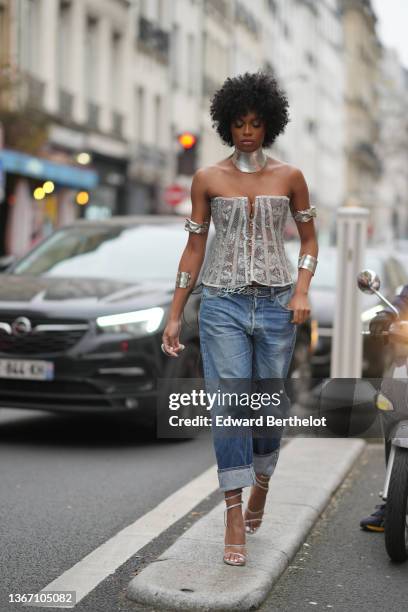 Didi Stone wears a silver large necklace, silver large shoulders bracelets, a silver laces buttoned / shoulder-off corset, blue faded denim large...