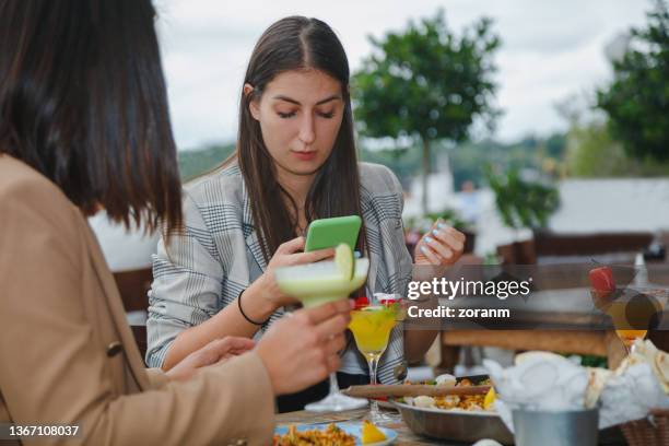 young woman sitting in outdoor restaurant with friend, using mobile phone - mexican food party stock pictures, royalty-free photos & images