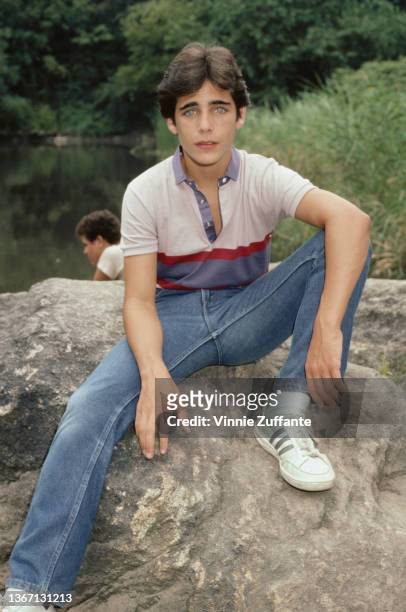 American actor Brian Bloom, wearing a grey polo shirt with a blue-and-burgundy stripe across the chest, and jeans, sitting on a rock, circa 1985.
