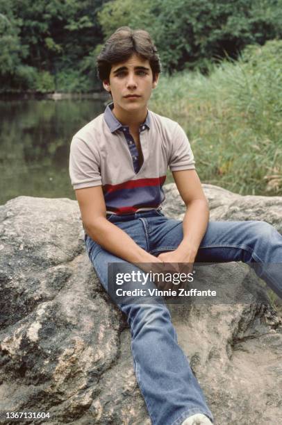 American actor Brian Bloom, wearing a grey polo shirt with a blue-and-burgundy stripe across the chest, and jeans, sitting on a rock, circa 1985.