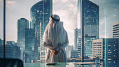 Successful Muslim Businessman in Traditional White Outfit Standing in His Modern Office Looking out of the Window on Big City with Skyscrapers. Successful Saudi, Emirati, Arab Businessman Concept.