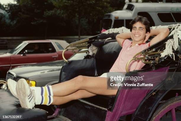 American actor Brian Bloom, wearing a red-and-white striped polo shirt with white shirts, reclining in a carriage, his hands behind his head, circa...