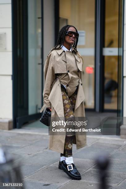 Guest wears black sunglasses, a white shirt, a beige long trench coat, black / yellow / white checkered print pattern legging pants, white socks,...