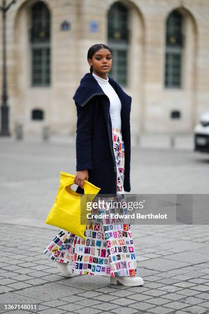 Guest wears a white ribbed turtleneck pullover, a navy blue long fluffy large collar coat, a yellow shiny leather large handbag, a white with...