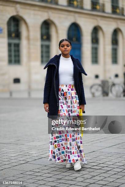 Guest wears a white ribbed turtleneck pullover, a navy blue long fluffy large collar coat, a yellow shiny leather large handbag, a white with...