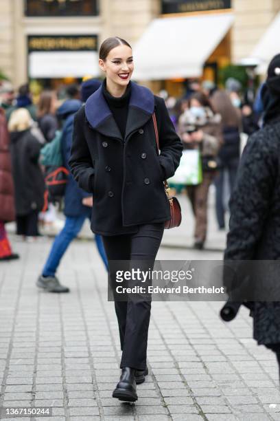 Model wears a black ribbed turtleneck pullover, a black with navy blue collar buttoned coat, a brown shiny leather shoulder bag, black denim...