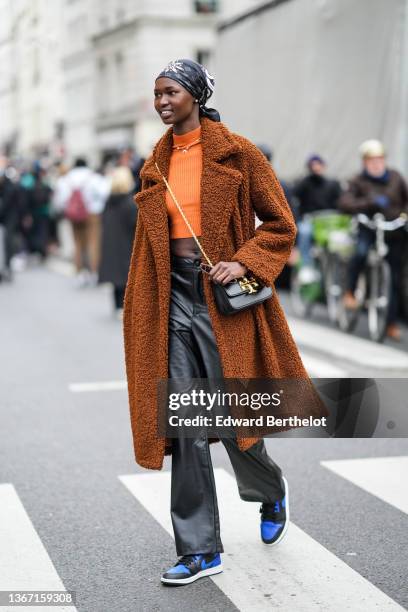 Model wears a black with white print pattern silk scarf, black sunglasses, an orange ribbed wool turtleneck pullover, a gold chains pendants...